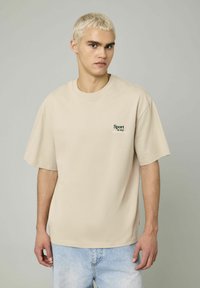 T-shirt en coton beige avec un col rond et des manches courtes, arborant un petit texte imprimé "Le sport est mon" en vert sur la poitrine.