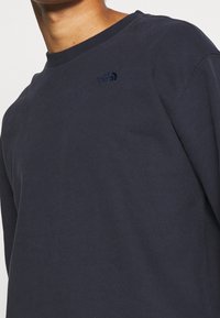 Muž s tmavě modrou mikinou s kulatým výstřihem, na níž je na hrudi malé vyšívané logo The North Face.