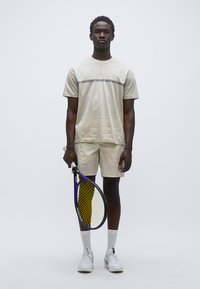 Αθλητικό πουκάμισο σε ανοιχτό μπεζ με λεπτή γκρι ρίγα, σετ με μπεζ σορτς. Κρατά ρακέτα τένις με κίτρινο και μοβ σχέδιο. Άσπρες κάλτσες και αθλητικά παπούτσια.