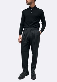 Polo nera a maniche lunghe con chiusura a cinque bottoni, abbinata a pantaloni neri plissettati a vita alta e scarpe nere con lacci.