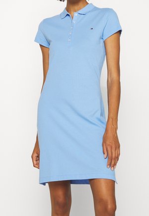 Woman wearing a light blue short-sleeve polo dress with a collar and button placket, featuring a small logo on the chest.