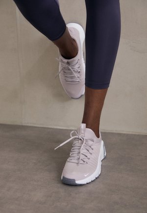 Personne portant des chaussures de course Nike rose clair et des leggings noirs, avançant sur un sol gris lisse.