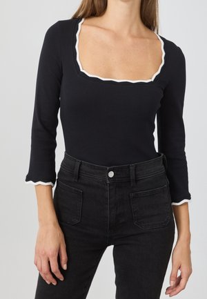 Frau trägt ein schwarzes langärmeliges Oberteil mit weißem, gewelltem Besatz am quadratischen Ausschnitt und an den Manschetten, kombiniert mit hoch taillierten schwarzen Jeans.