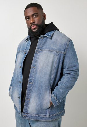 Brodaty mężczyzna w czarnej, warstwowej bluzie z kapturem i jasnoniebieskiej dżinsowej kurtce, ze złożonymi rękami w kieszeniach, stojący na jednolitym jasnym tle.