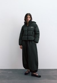 Mladá žena v čiernej vatovanej bunde, voľných čiernych nohaviciach a čiernych balerínach stojaca pri jednoliacej bielej stene.