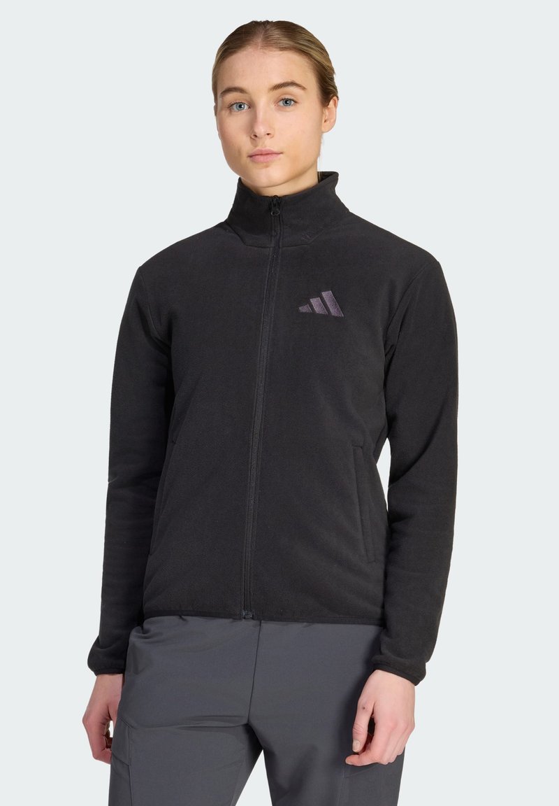 Νεαρή γυναίκα φοράει μαύρο fleece μπουφάν με φερμουάρ και μικρό λογότυπο στο στήθος και γκρι παντελόνι, στέκεται μπροστά σε ένα απλό φόντο.