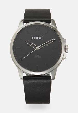 Relógio analógico preto com caixa redonda em aço inoxidável, bracelete de pele pretas texturizadas e mostrador preto com ponteiros e marcadores prateados de horas/minutos.