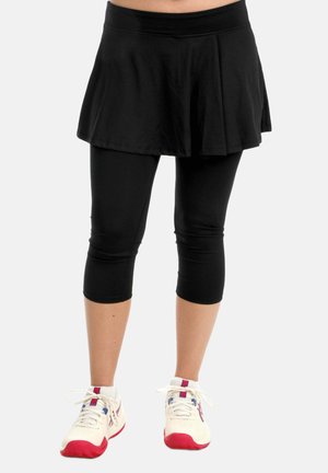 Femme portant une jupe-short noire avec des leggings intégrés et des chaussures de sport blanches à semelles rouges, debout devant un fond uni.