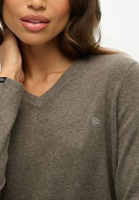 Brązowy sweter z dekoltem w serek wykonany z miękkiego materiału, z małym haftowanym logiem na piersi i ściągaczami przy rękawach.