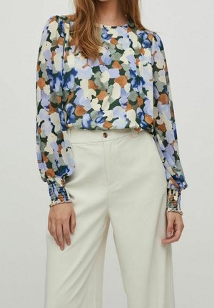 Vrouw draagt een blouse met lange mouwen in een bloemmotief van blauw, groen, bruin en wit, gecombineerd met een broek in off-white kleur met een hoge taille en wijde pijpen.