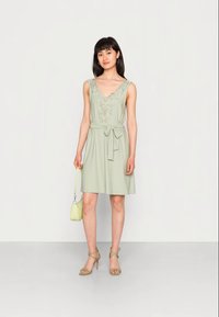 Vestido sem mangas verde-claro com decote em V e detalhes bordados, atado na cintura. Combinado com sapatos de salto verdes e uma pequena bolsa.