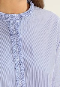 Tyrkysovo-bílá pruhovaná blůza s volánkovým límcem a légou. Tkanina působí lehce, s vertikálními pruhy a jemnými detaily.