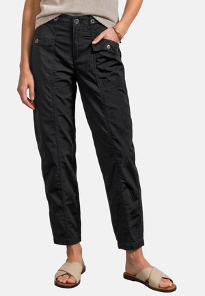 Personne portant un pantalon cargo noir avec des poches boutonnées et des sandales beiges à brides croisées sur un fond clair.