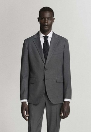 Uomo con abito grigio, camicia bianca e cravatta nera, in piedi davanti a uno sfondo chiaro e semplice, rivolto in avanti.