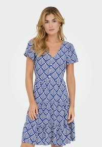 Femme avec de longs cheveux blonds portant une robe V-neck à motifs bleus et blancs, avec des manches courtes et amples et des boutons à l'avant.