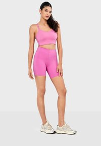 Femme portant un crop top sportif rose et un short de cycliste taille haute assorti, chaussée de chaussures de course blanches, sur un fond clair uni.