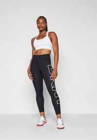 Black leggings with bold white lettering on the side, paired with a white sports bra. Smooth fabric and athletic fit. Sneakers with orange accents.
