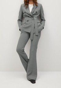 Femme portant un blazer gris cintré à double boutonnage avec ceinture et un pantalon évasé assorti, debout les mains dans les poches.