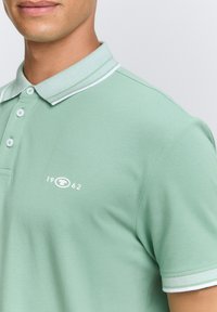 Hellgrünes Poloshirt aus strukturiertem Stoff, mit einem gerippten Kragen und weißen Akzenten sowie einem kleinen Logo mit dem Jahr 1962.