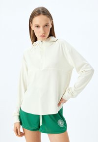 Sudadera blanca con cremallera y capucha, con mangas largas y material suave; combinada con pantalones cortos deportivos verdes con estampado de logo.