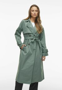 Trench coat verde lungo realizzato in un materiale morbido, con frontale doppiopetto, vita con cintura e ampie tasche laterali.