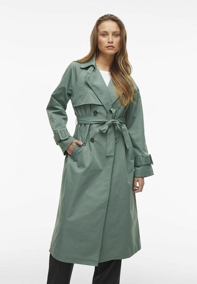 Trench coat verde lungo realizzato in un materiale morbido, con frontale doppiopetto, vita con cintura e ampie tasche laterali.