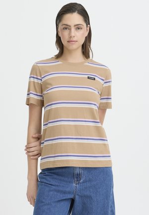 Frau mit dunklen Haaren trägt ein beiges T-Shirt mit lila-weißen horizontalen Streifen und blaue Jeans, steht vor weißem Hintergrund.