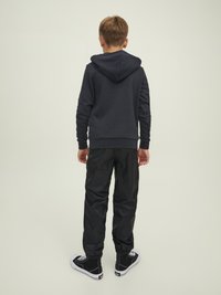 Sudadera negra con un corte relajado, combinada con pantalones deportivos negros y brillantes y zapatillas negras. Ropa deportiva informal con una textura ligeramente marcada.