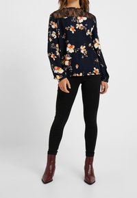 Blusa floreale con base blu navy, maniche lunghe e dettagli in pizzo nero sul collo. Indossata con pantaloni neri e stivaletti borgogna.