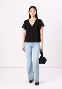Black polka dot top with sheer ruffled sleeves, V-neck design, paired with light blue flared jeans and a black floral bag. Black flats.