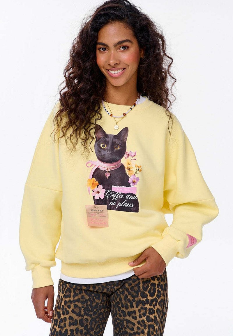 Mladá žena s kudrnatými tmavými vlasy má na sobě žlutou mikinu s černou kočkou, květinami a textem "Káva a žádné plány", doplněnou leopardími kalhotami.