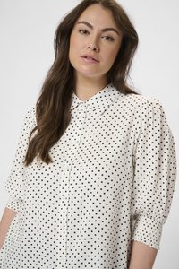 Blouse blanche avec des pois noirs, devant à boutons, manches courtes bouffantes avec des poignets élastiques, texture lisse et col classique.