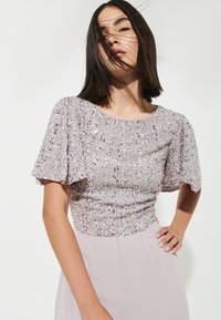 Top in lavanda con scollatura rotonda, maniche corte a volant e paillettes decorative che creano un motivo testurizzato e scintillante. Tessuto liscio e leggero sul fondo.