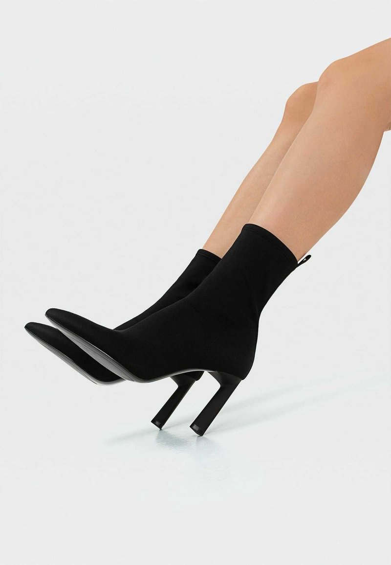 Jambes portant des bottes chaussettes extensibles noires avec des bouts pointus et des talons blocs inclinés sur un fond uni.