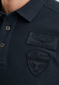Marineblaues Poloshirt aus strukturiertem Stoff, ausgestattet mit zwei Knöpfen und zwei geprägten Aufnähern mit Logo und Markendetails.