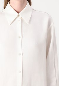 Camicia bianca con bottoni, dal tessuto morbido e liscio. Presenta un colletto classico e cuciture decorative lungo le cuciture delle spalle.