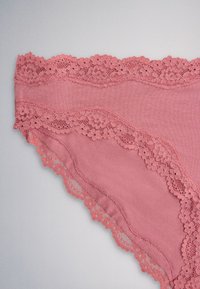 Pink dameundertøj med blomsterblondekant langs taljen og benåbningerne på en neutral baggrund.