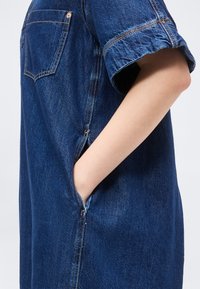 Personne portant une robe en denim bleue avec manches courtes retroussées, main posée de manière décontractée dans la poche latérale, sur un fond blanc uni.