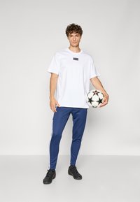 Hvit t-skjorte med rund hals og logo-patch, kombinert med blå smale joggebukser med hvite striper og svarte joggesko, holder en fotball.