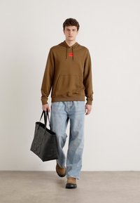 Brun hettegenser med frontlomme, sammen med lyseblå jeans. Svart totebag med pregede detaljer. Brune sko.