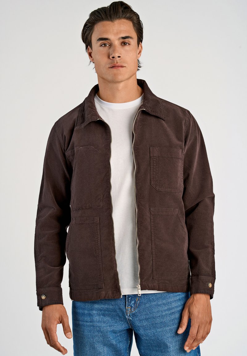 Chaqueta de lona marrón con cremallera en la parte delantera, dos bolsillos en el pecho y puños abotonados, usada sobre una camiseta blanca y combinada con unos vaqueros azules.