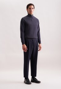 Uomo in piedi, indossando un maglione grigio scuro con zip, pantaloni a pieghe blu navy, calzini neri e scarpe eleganti in pelle nera su uno sfondo chiaro.
