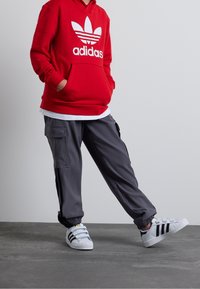 Röd huvtröja med vitt Adidas-logotyp, grå cargobyxor med fickor och vita sneakers med svart och grå randdesign.