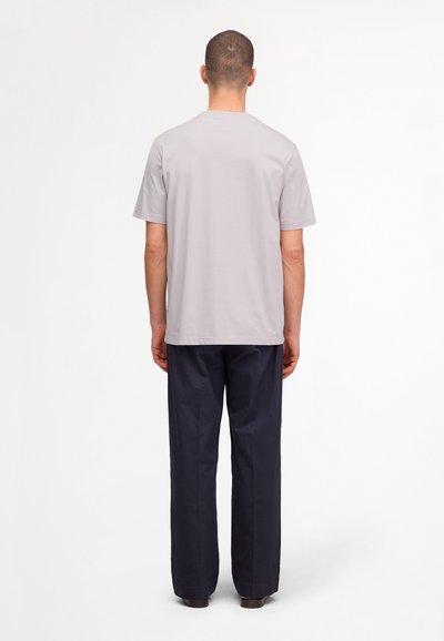 Homme debout de dos, portant un t-shirt gris clair à manches courtes et un pantalon droit bleu marine foncé sur fond blanc.