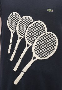 Haut sans manches en tricot marine avec un graphique blanc représentant quatre raquettes de tennis sur un motif en filet. Petit logo en haut à gauche.