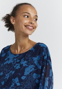 Blusa blu navy con motivo floreale in varie tonalità di blu. Tessuto morbido e fluido con scollatura rotonda e maniche trasparenti.