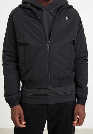 Hombre con chaqueta negra con cremallera sobre sudadera negra con capucha y pantalones negros, con el logo "cK" en el pecho izquierdo de la chaqueta.