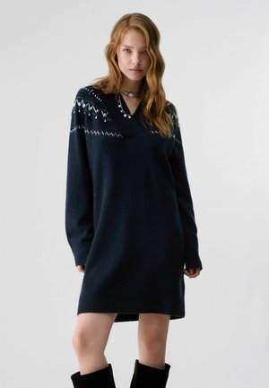 Jeune femme aux cheveux ondulés portant une robe-pull en tricot bleu foncé avec des motifs géométriques blancs et des bottes noires, debout devant un fond uni.