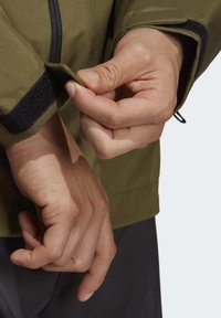 Olivgrön jacka med justering av ärmslut med kardborre, som visar strukturerad tyg och en elegant design. Händer som justerar ärmslutsdetaljen.
