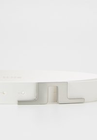 Vit läderbälte med en metallisk silverfärgad rektangulär spänne med geometrisk design. Inkluderar jämnt fördelade hål för justering.